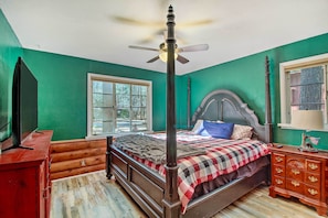Quarto com cama de dossel, cômoda de madeira, televisão e janela com vista para árvores.