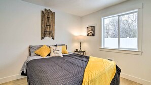 A bedroom with a large bed, a nightstand, a lamp, a window with a view of trees, and a wall with a decorative piece. - Cozy Blackfoot Apartment: Pets Welcome! (Blackfoot)