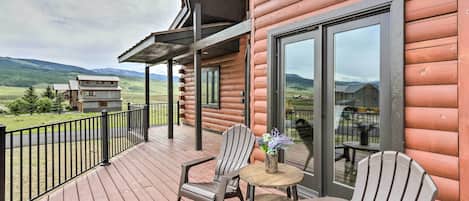 A wooden cabin with a deck, two Adirondack chairs, a small round table, and a large sliding glass door.