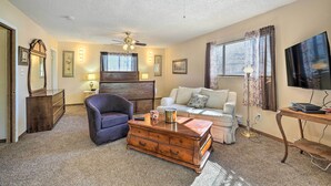 Un salon avec un ventilateur de plafond, une commode en bois, un fauteuil sombre, une table basse en bois, un canapé blanc et un téléviseur fixé au mur.
