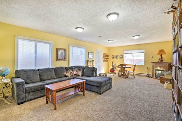 A living room with a sectional sofa, a coffee table, a fireplace, a bookshelf, and a dining area with chairs and a table. - Pet-friendly Duplex - Near Skyline Drive! (Canon City)