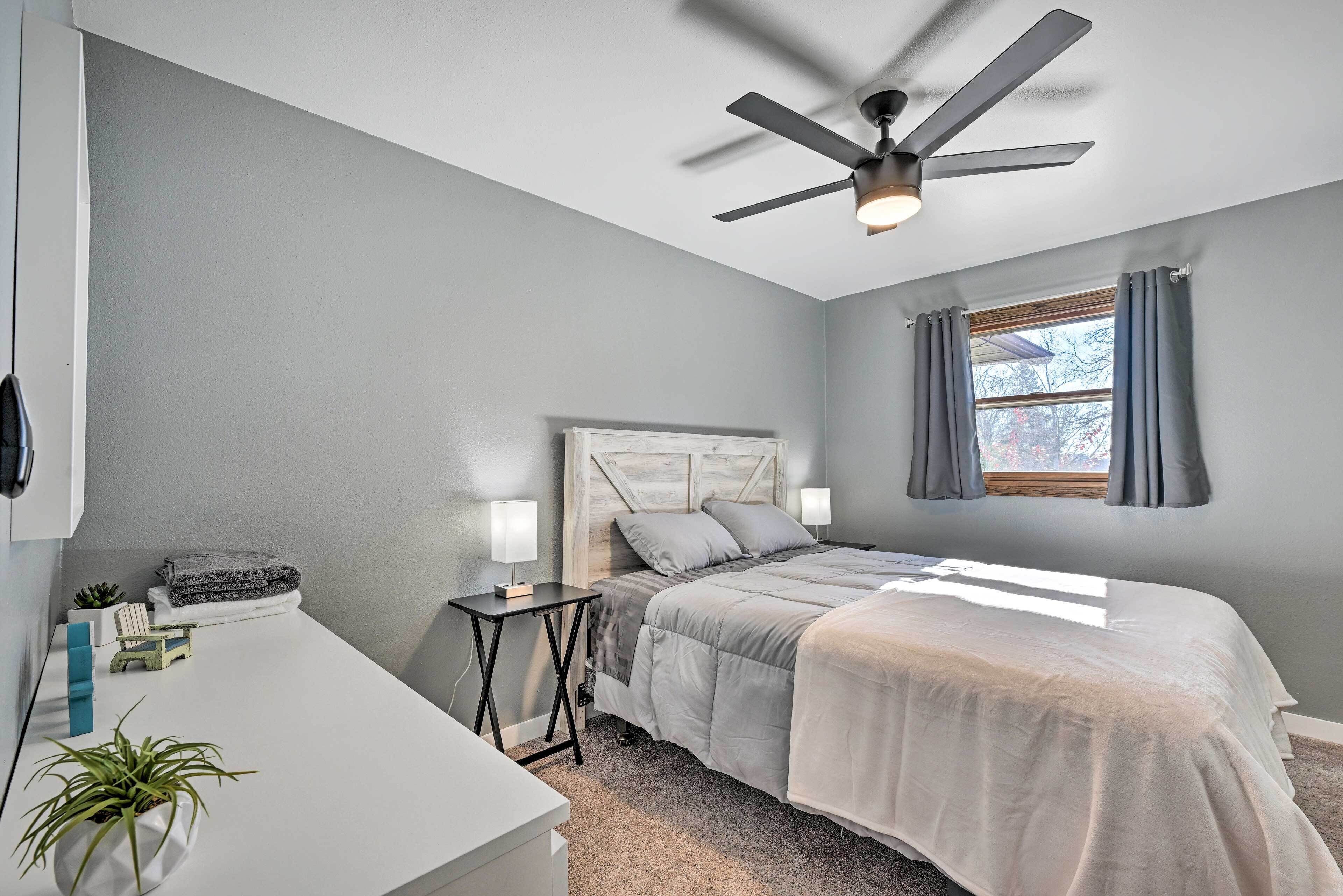 Une chambre à coucher avec un lit, des lampes de chevet, un ventilateur de plafond, une fenêtre avec des rideaux et une petite plante posée sur une table de chevet.