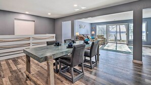 A spacious dining area with a long wooden table and modern chairs, a white railing, and a view of a deck through large windows. - Charming Wausau Cottage: On-site Lake Access! (Wausau)