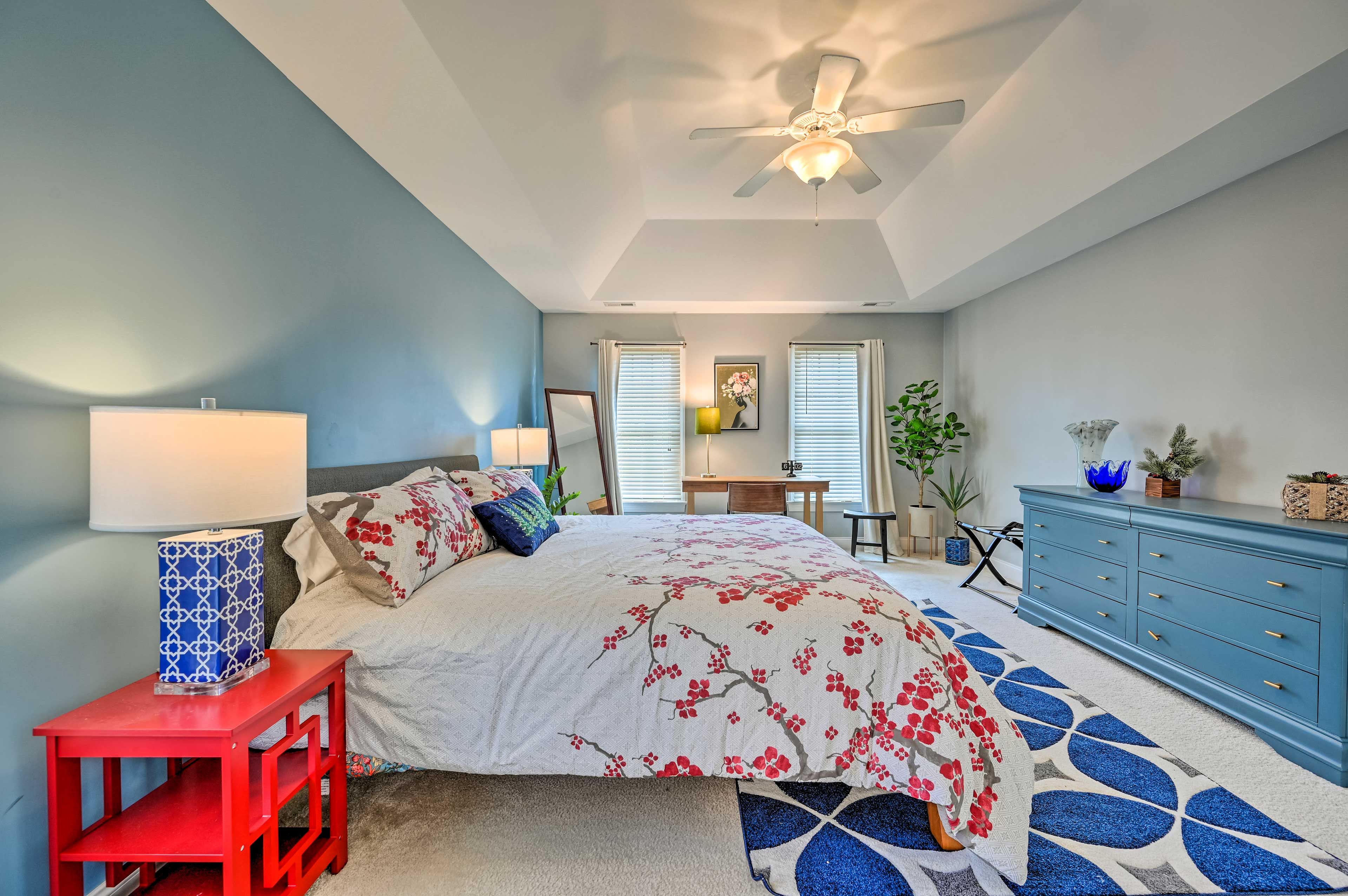 A bedroom with a bed, a red nightstand, a blue dresser, a ceiling fan, and a desk with a chair.