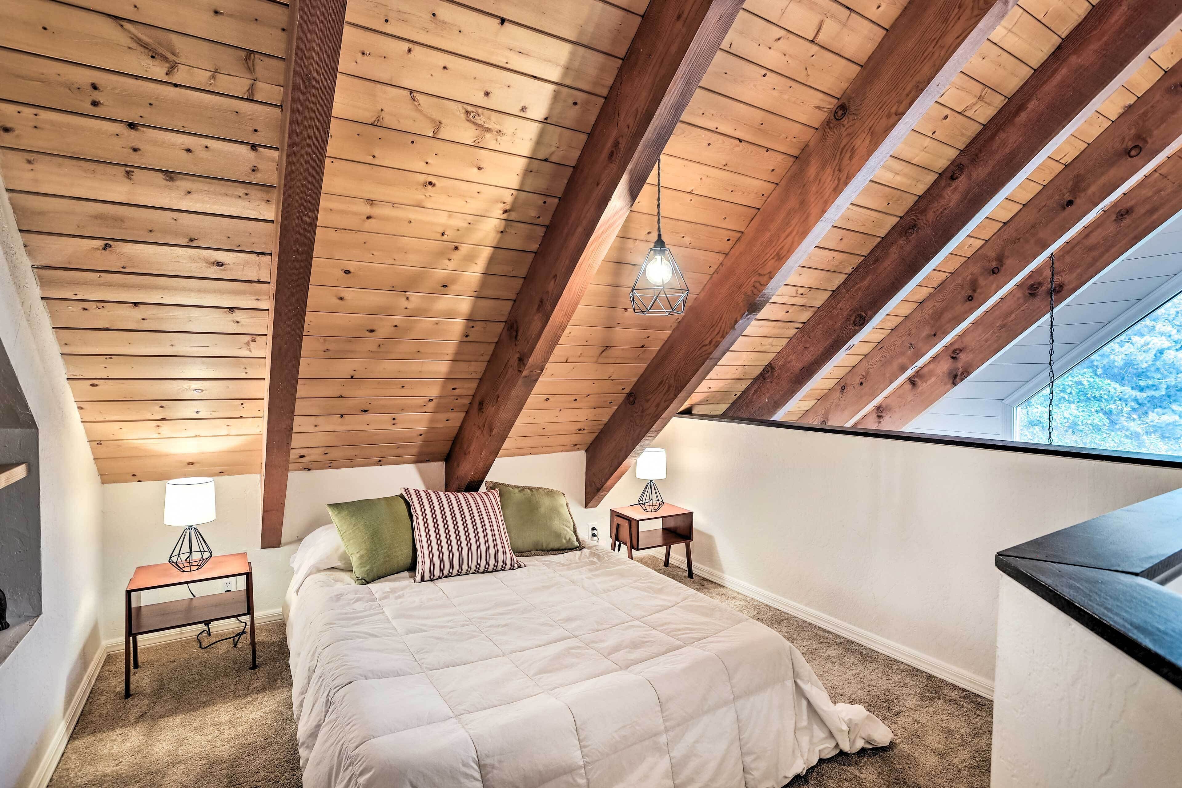 A bedroom with a sloped ceiling, wooden beams, a bed with pillows, bedside tables with lamps, and a view through a window.
