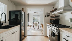 Une cuisine moderne avec des meubles blancs, un réfrigérateur noir, une cuisinière et un four à micro-ondes. On aperçoit un coin repas avec une table et des chaises à travers une porte ouverte.