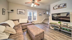 A living room with a sectional sofa, ottoman, ceiling fan, mounted TV, and framed pictures on the walls. - Charming Kellogg Home Near Outdoor Activities (Kellogg)