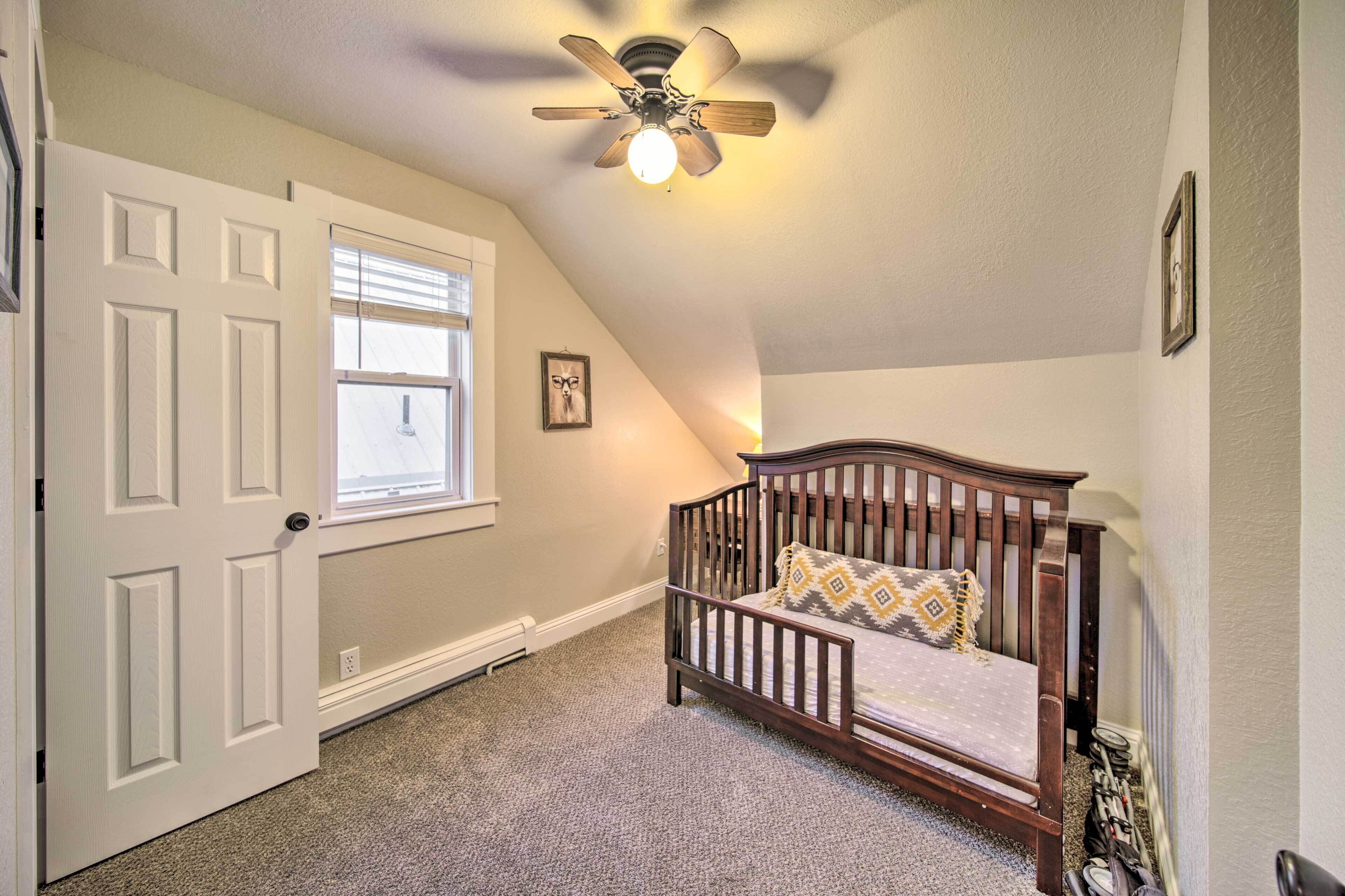 A bedroom with a sloped ceiling, a wooden crib, a ceiling fan, and a window with blinds.