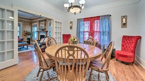 A dining room with a round wooden table, six wooden chairs, a red upholstered chair, a chandelier, and a window with red curtains. - Burgaw House w/ Large Covered Porch & Swing! (Burgaw)