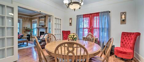 Une salle à manger avec une table ronde en bois, six chaises en bois, un fauteuil recouvert de tissu rouge, un lustre et une fenêtre ornée de rideaux rouges.