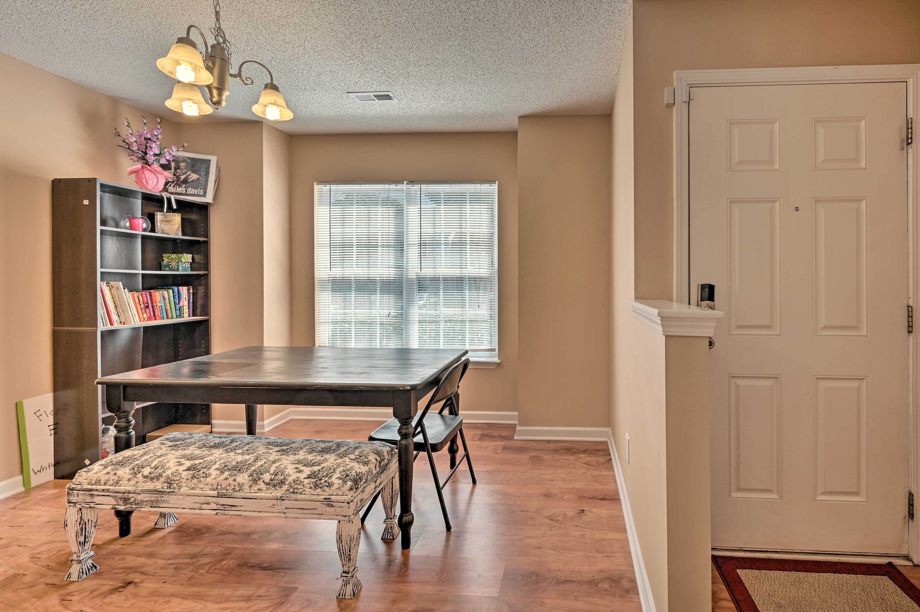A dining area with a wooden table, chairs, and a bench. There is a bookshelf with books and decorative items. A window with blinds is visible in the background.