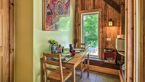Un coin repas en bois avec une table dressée pour quatre personnes, un tableau abstrait aux couleurs vives accroché au mur et une fenêtre donnant sur un paysage verdoyant.