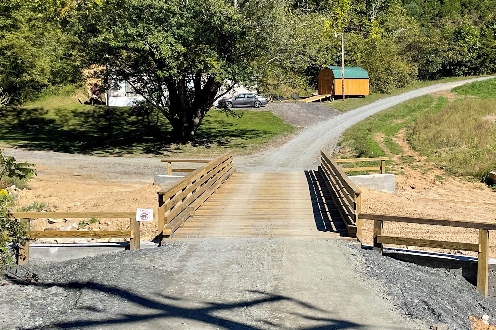 A wooden bridge leading to a dirt road with a small building in the background.