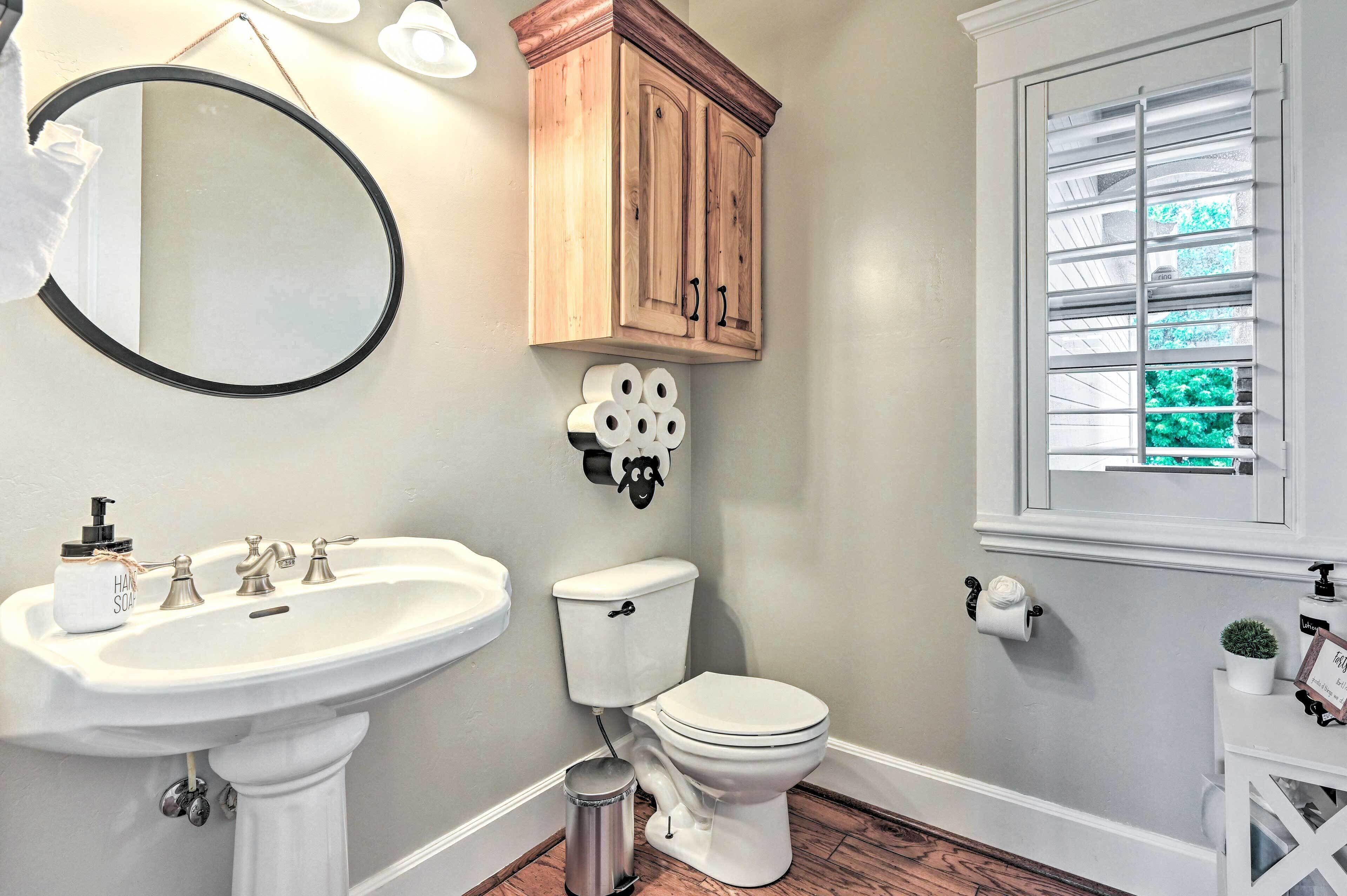 A bathroom with a white sink, toilet, and mirror. There is a wooden cabinet above the toilet and a window with blinds.