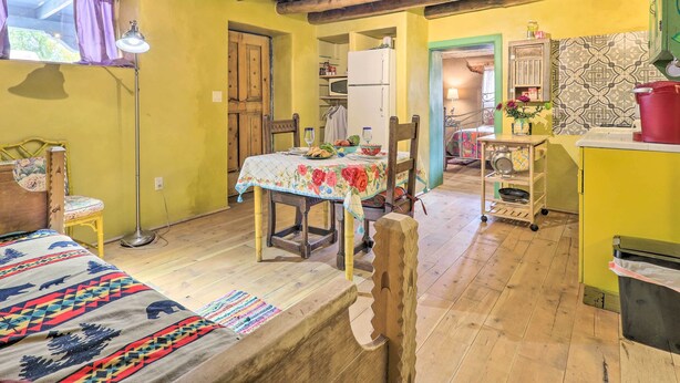 A cozy room with a bed, a dining table with a floral tablecloth, wooden chairs, a wooden floor, a ceiling with exposed beams, a window with curtains, a lamp, and a refrigerator in the background. - Artsy Santa Cruz Apartment w/ Shared Fire Pit! (Santa Cruz)