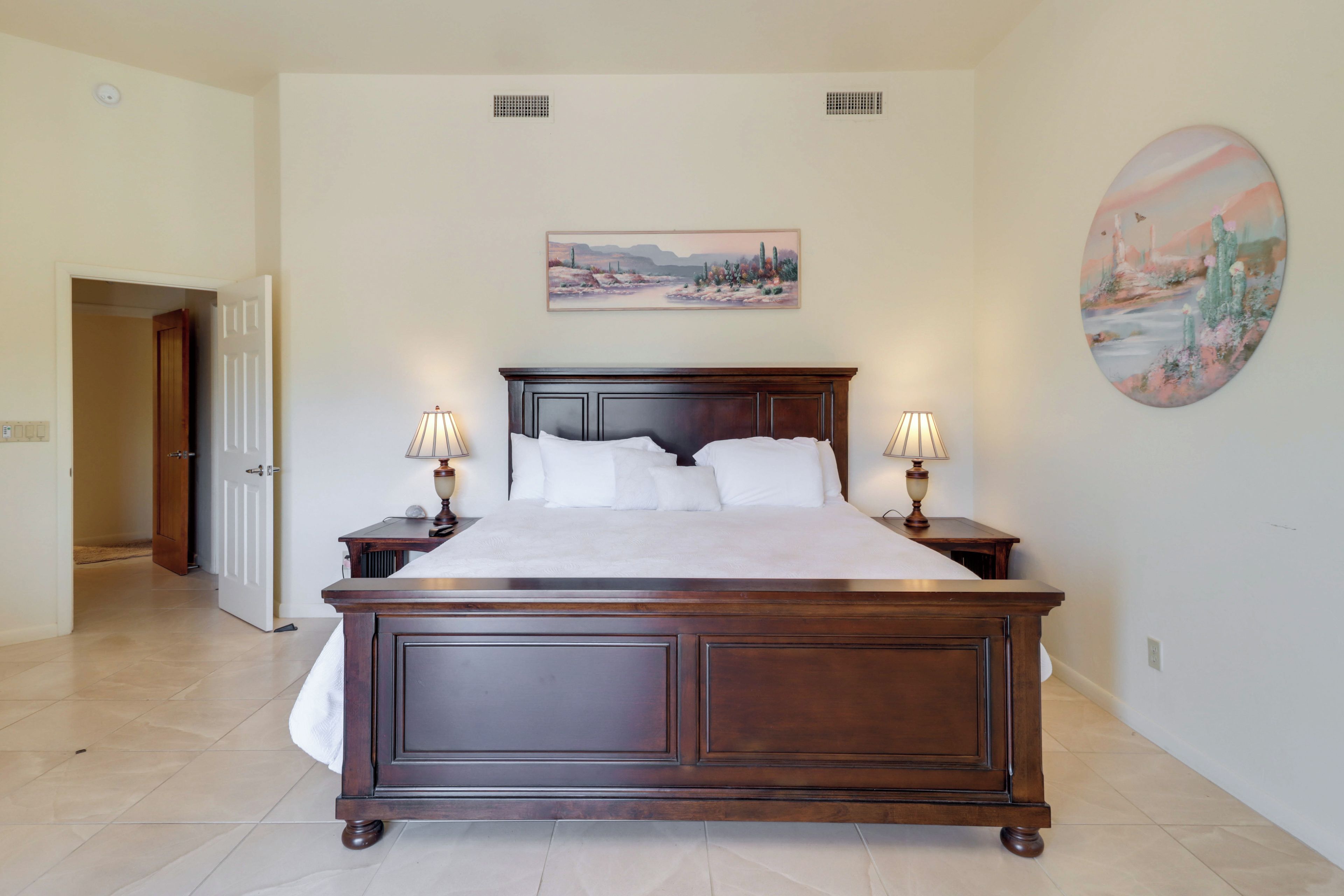 A large bed with a dark wooden frame, two bedside tables with lamps, a painting on the wall, and a door leading to another room.
