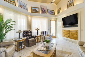 A spacious living room with a leather sofa, a coffee table, a TV mounted on the wall, and a large window with blinds.