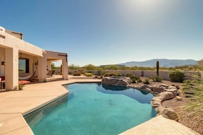 A modern outdoor pool area with a clear blue pool, surrounded by rocks and desert plants, and a view of mountains in the background.