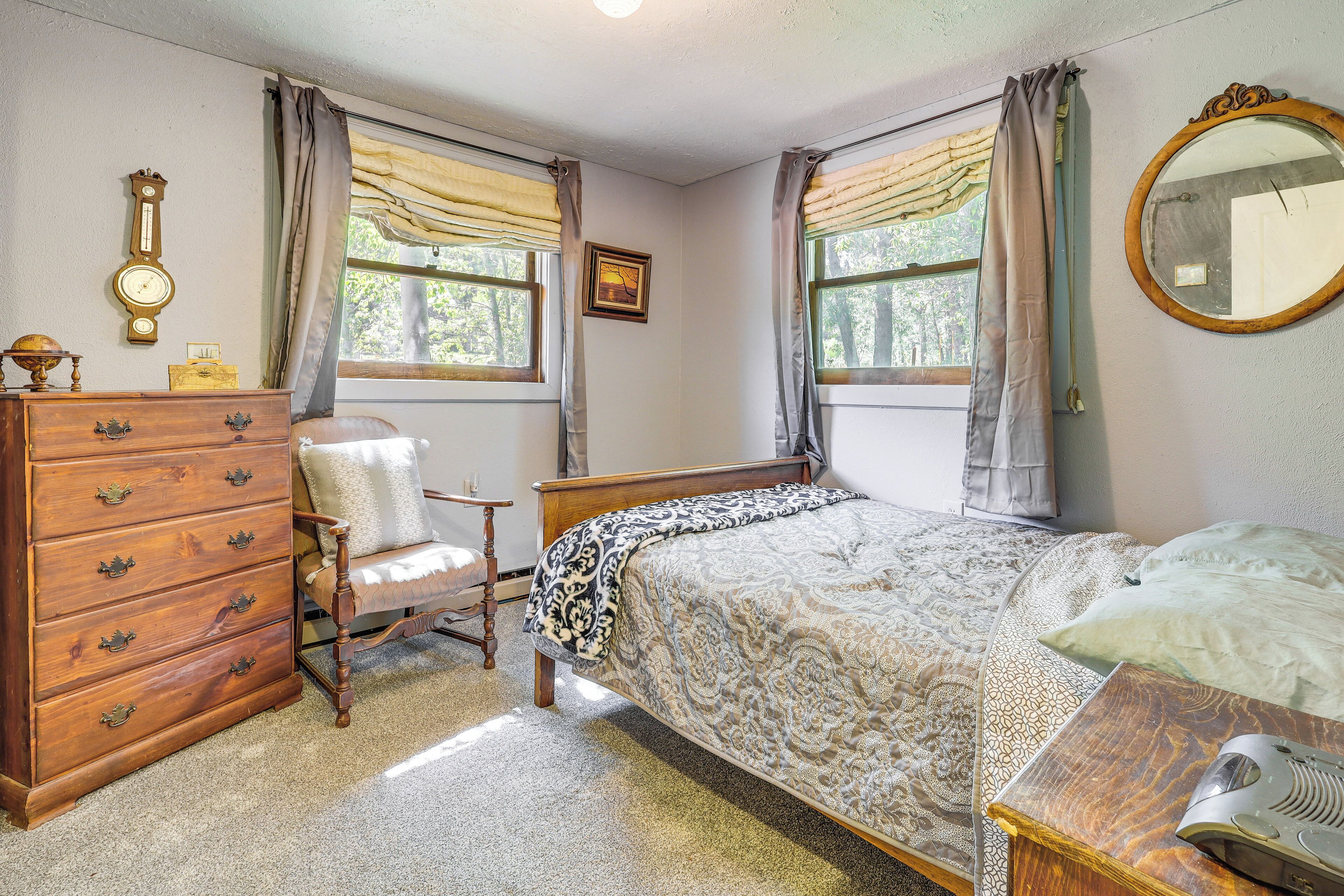 A bedroom with a wooden dresser, a chair, a bed with patterned bedding, a mirror, and two windows with curtains.
