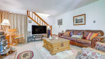 Un salon comprenant un coffre en bois, un canapé en cuir marron, un fauteuil à bascule en bois, un téléviseur et un escalier.