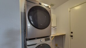 A compact laundry area with a stacked washer and dryer, a white door, and a small shelf with laundry baskets. - Gilbert Home w/ Pool < 5 Mi to Downtown! (Gilbert)