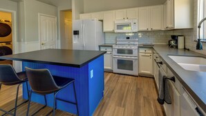 A modern kitchen with white cabinets, a blue island with two bar stools, a refrigerator, microwave, oven, and dishwasher. - Gilbert Home w/ Pool < 5 Mi to Downtown! (Gilbert)