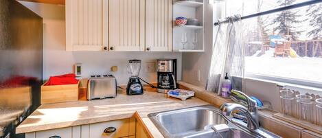A compact kitchen with white cabinets, a sink, a toaster, a coffee maker, and a window with a view of trees.