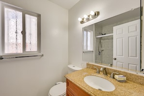 Une salle de bain avec des toilettes blanches, un lavabo doté d’un plan de travail en granit, un miroir avec éclairage intégré et une porte de douche.