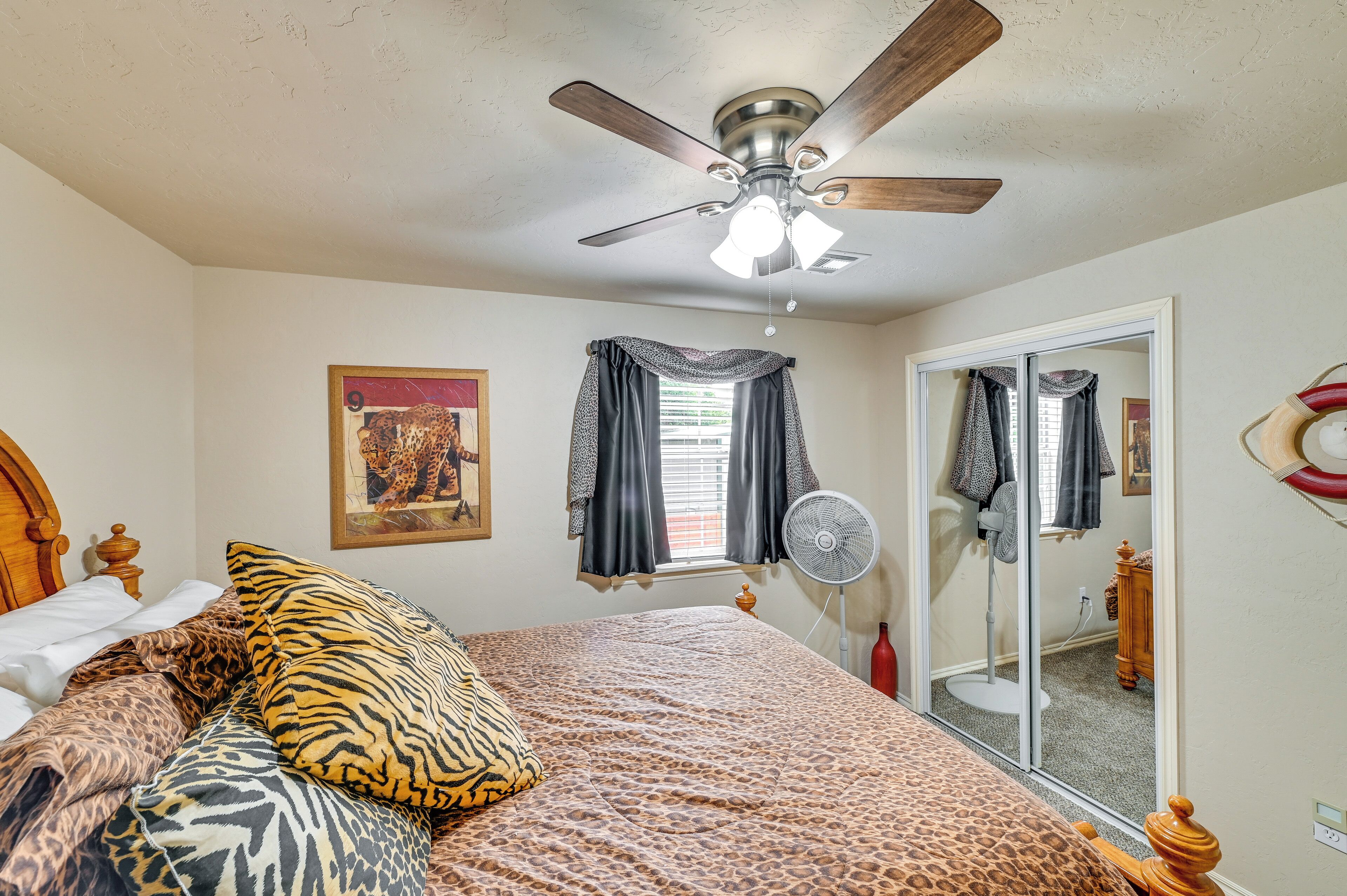 A bedroom with a bed, a ceiling fan, a window with curtains, a mirror, and a fan on the floor.