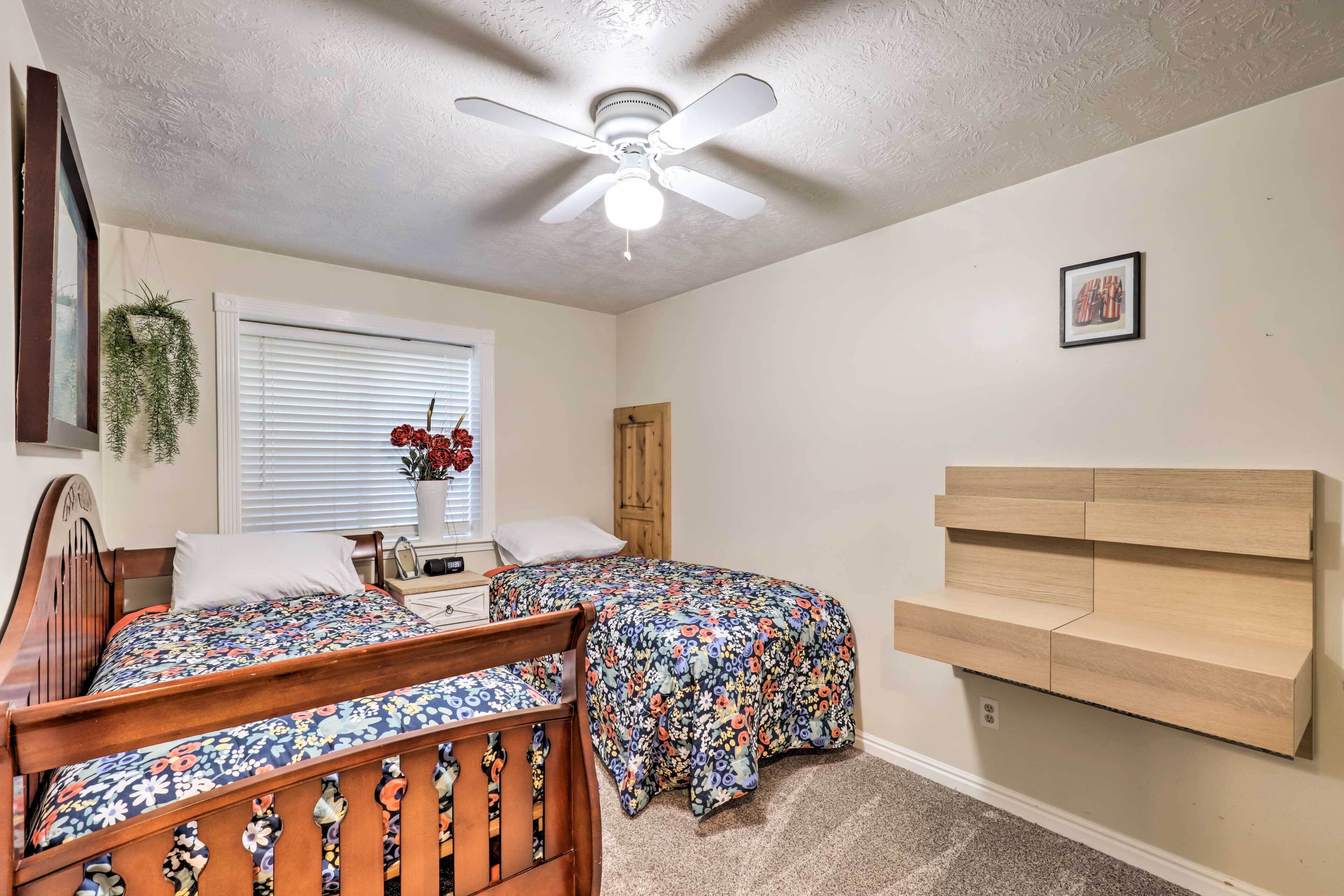 A bedroom with two beds, a ceiling fan, a wooden headboard, a small bedside table, and a window with blinds.