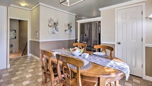 Un comedor con una mesa de madera, sillas y un tazón de fruta. Se observa un refrigerador y una puerta al fondo.