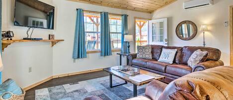 A cozy living room with a wooden ceiling, a flat-screen TV, a sofa set, a coffee table, and a large window with curtains.