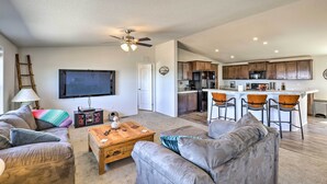 A spacious living room with a gray leather couch, a wooden coffee table, and a ceiling fan. A kitchen with wooden cabinets and a bar area is visible in the background. - Desert Escape Off I-10 w/ Over 1 Enclosed Acre! (Benson)