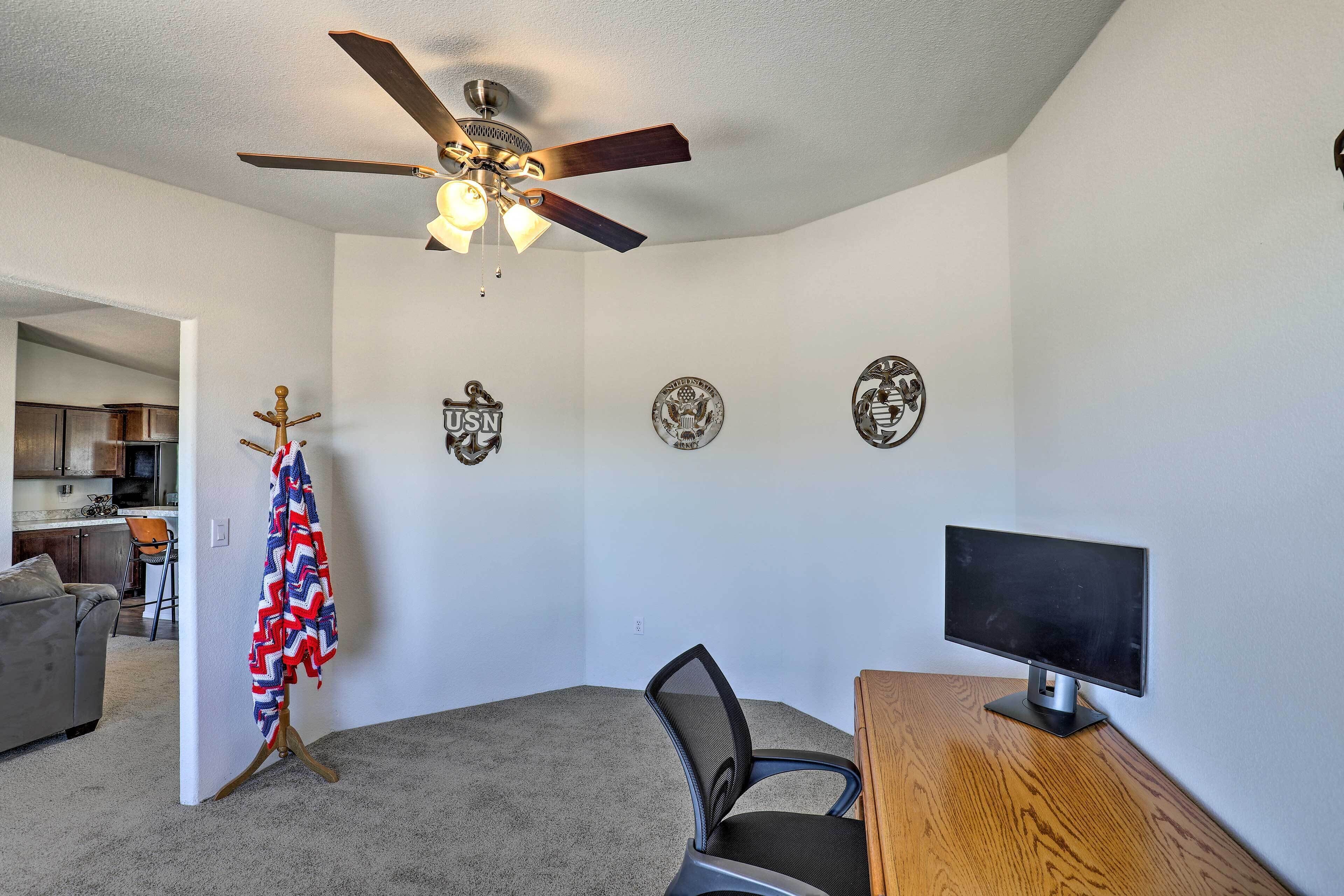 A room with a ceiling fan, a desk with a computer, a chair, and a coat rack with a flag.