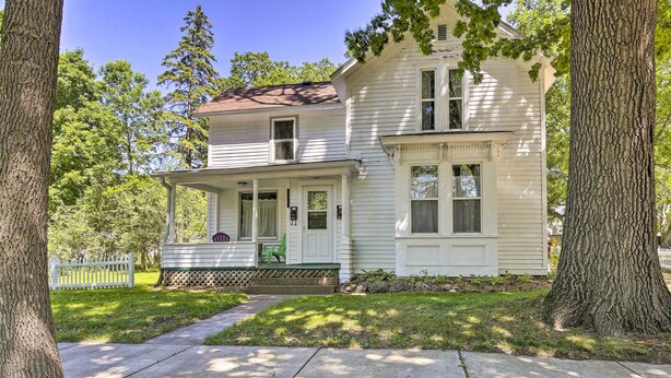 A two-story white house with a front porch, a small porch area with chairs, and a white picket fence.
