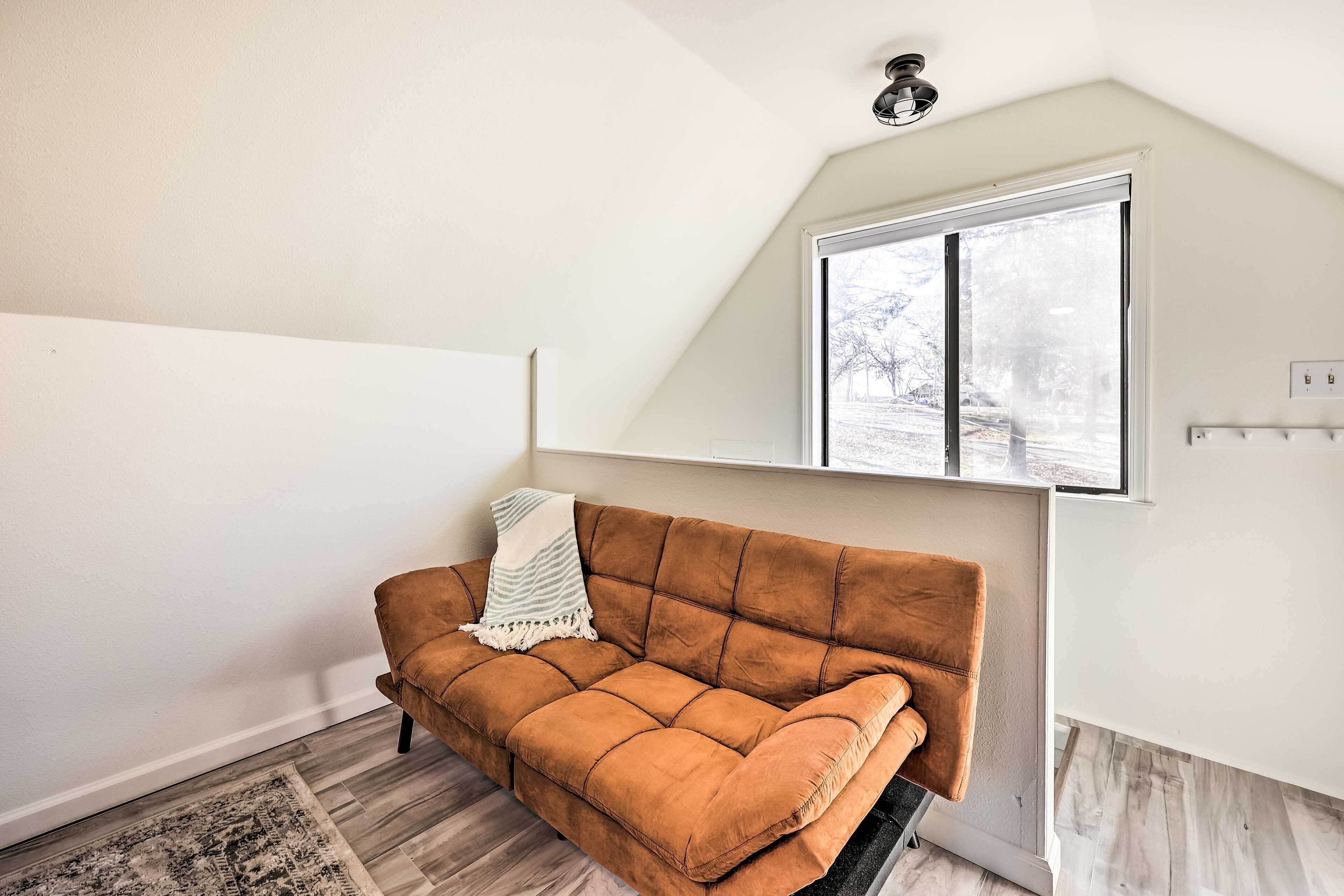 Ein Zimmer mit einem braunen Sofa, einem gemusterten Teppich, einem Fenster mit Blick auf Bäume und einer Deckenleuchte.