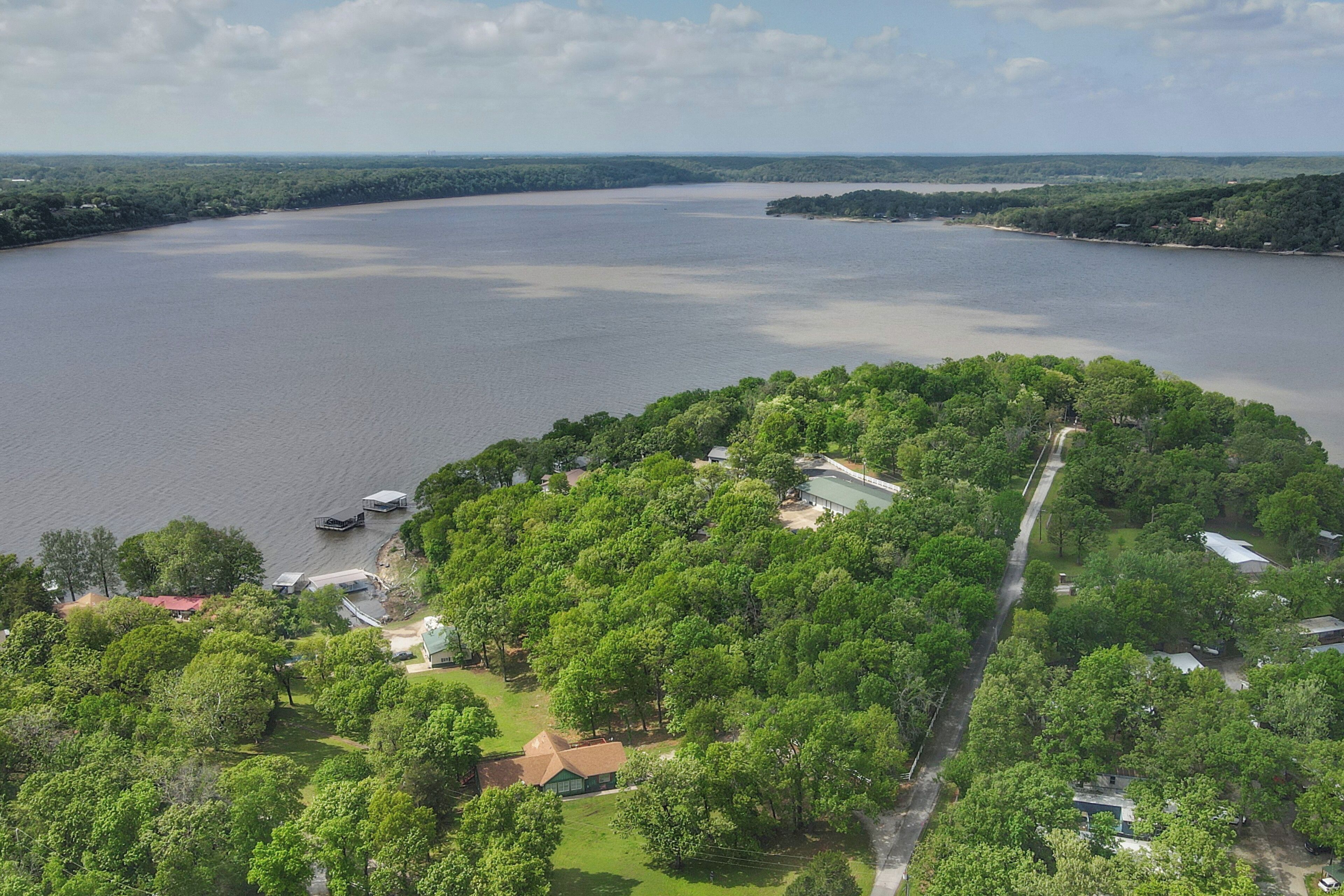 An aerial view of a lakeside property with dense greenery, a road, and a few buildings.