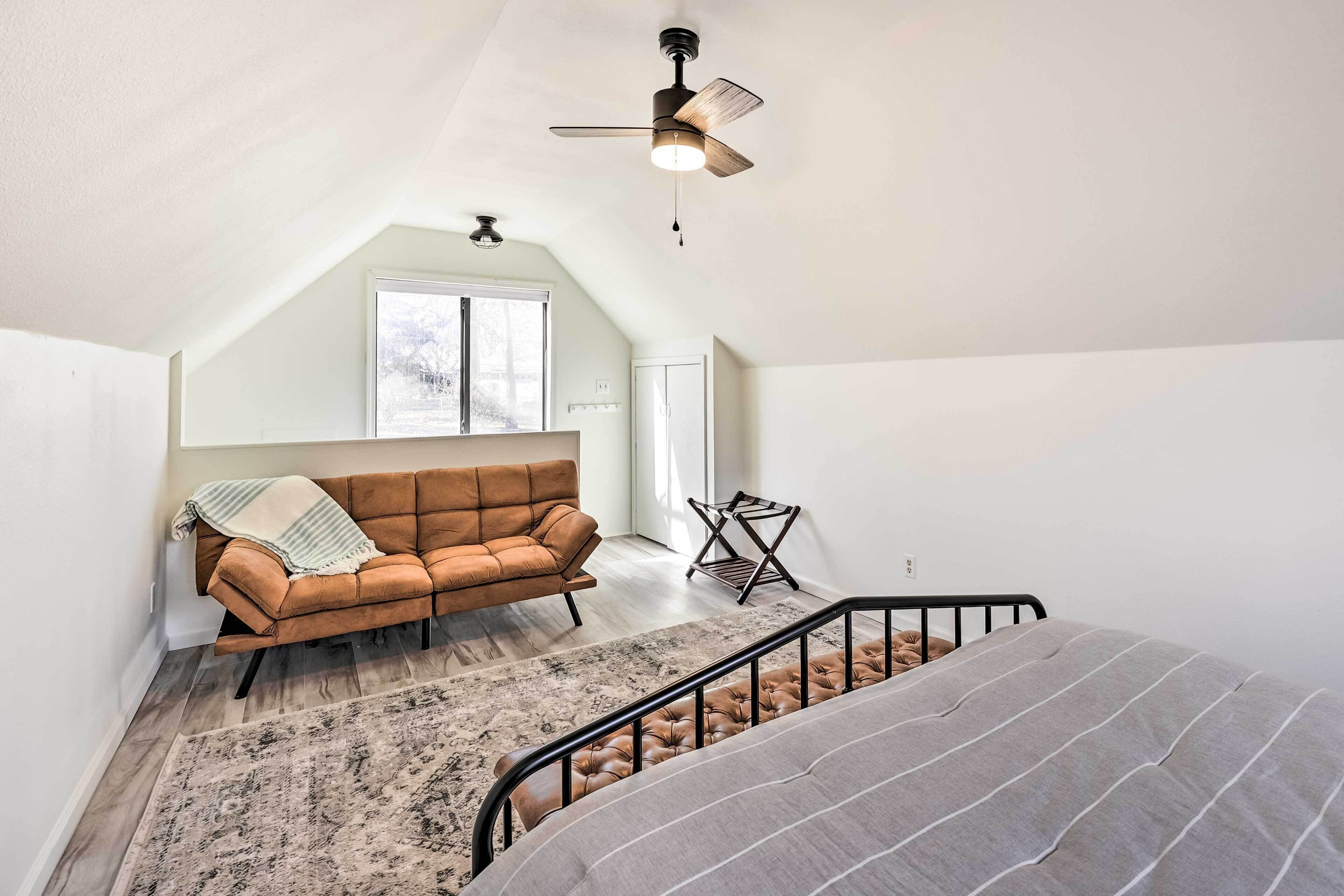 A loft bedroom with a sloped ceiling, a brown leather sofa, a patterned rug, a bed with striped bedding, and a ceiling fan.