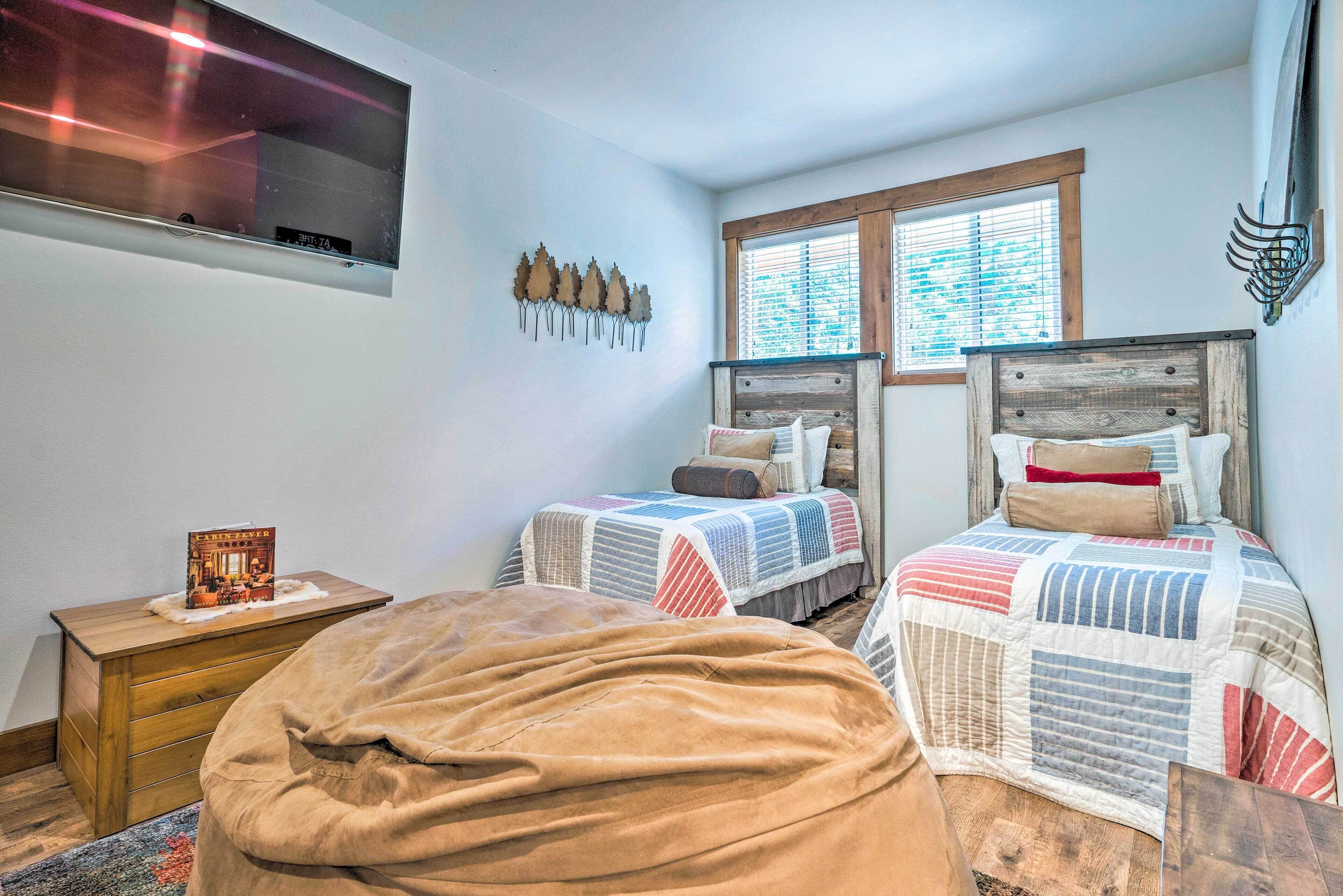 Une chambre avec deux lits, une commode en bois, un téléviseur fixé au mur et une fenêtre avec des stores.
