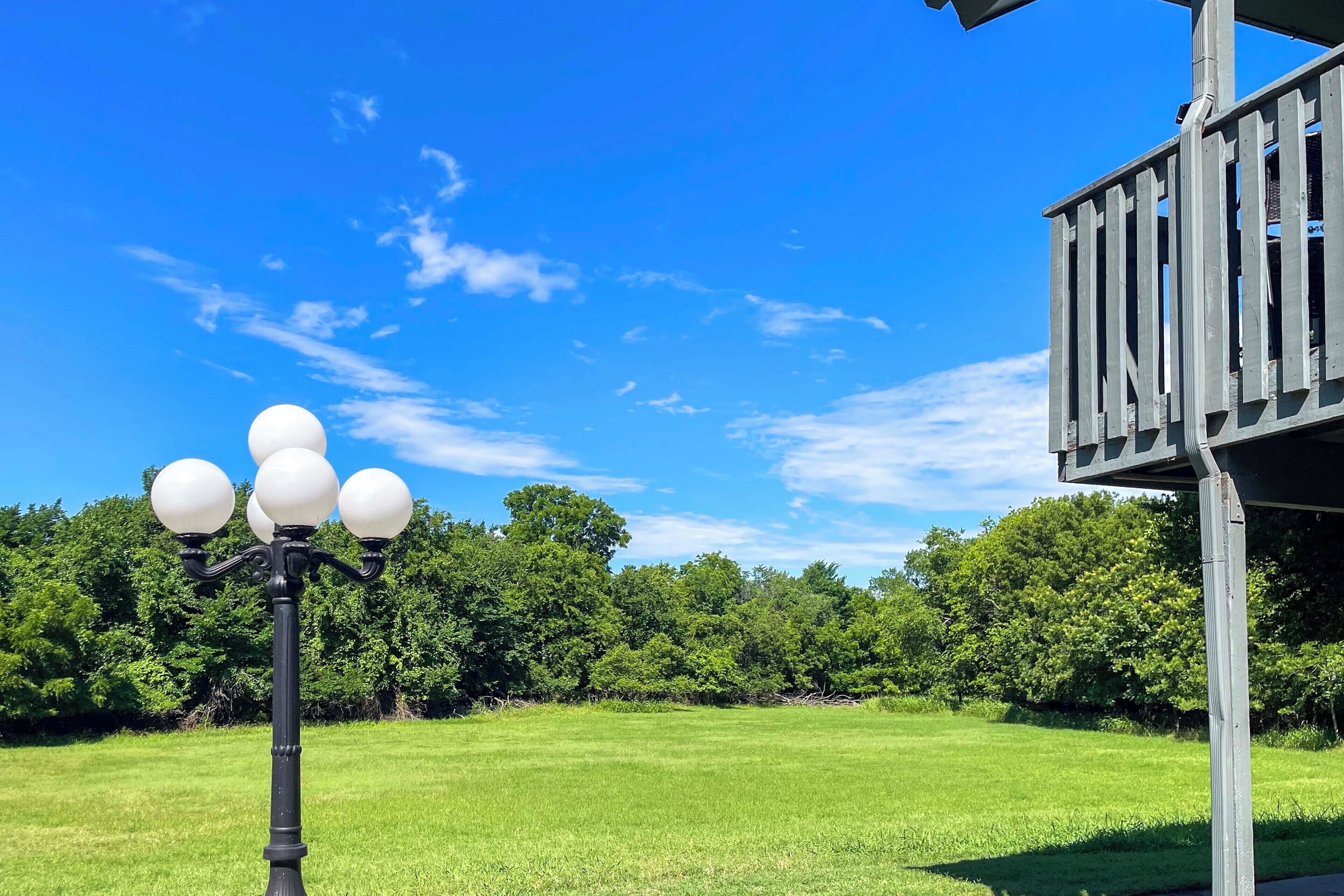A black street lamp with four globes, a wooden deck, and a grassy field with trees in the background.