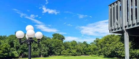 A black street lamp with four globes, a wooden deck, and a grassy field with trees in the background.
