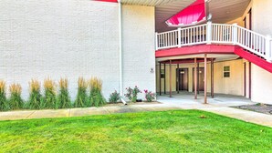 A building with a red and white exterior, a covered outdoor area with a white railing, and a small garden with grass and plants. - Cozy Pigeon Getaway, Close to Public Beaches! (Pigeon)