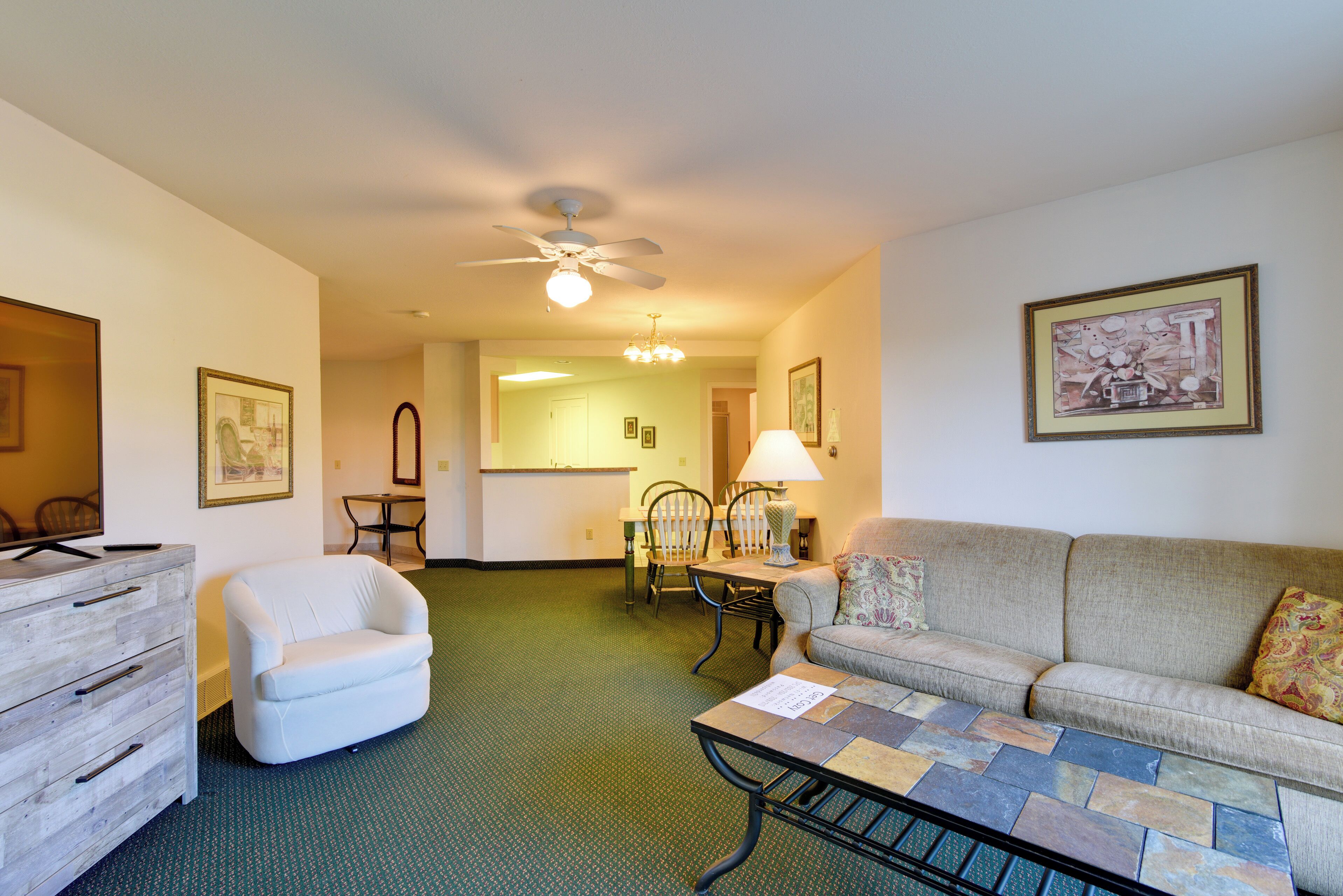 A hotel room with a green carpet, a white armchair, a beige sofa, a coffee table, a wooden dresser, a ceiling fan, and framed artwork on the walls.