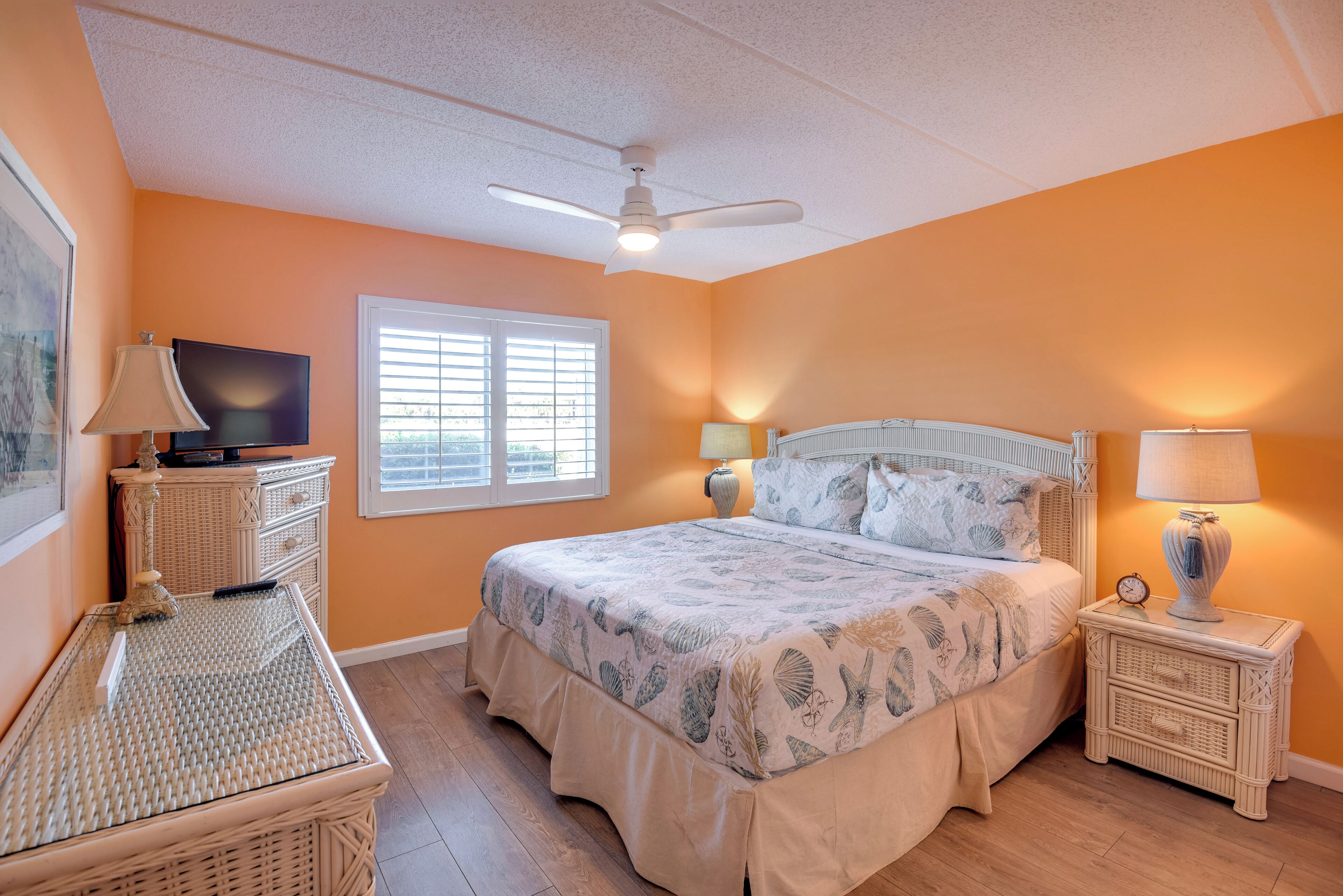 A bedroom with a bed, a television, a window, a ceiling fan, and a nightstand with a lamp.