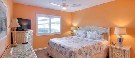 A bedroom with a bed, a television, a window, a ceiling fan, and a nightstand with a lamp.