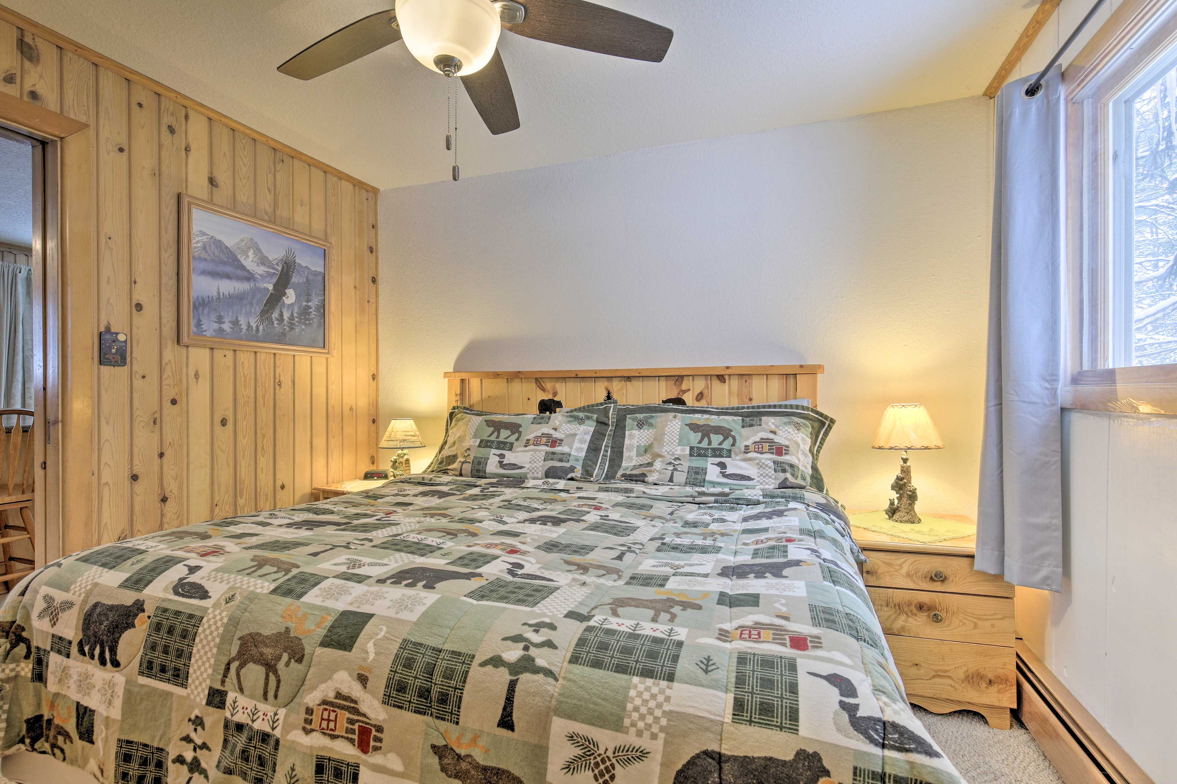 Un dormitorio con pared de paneles de madera, ventilador de techo, cama con colcha estampada de animales, dos mesitas de noche con lámparas y una ventana con cortinas.