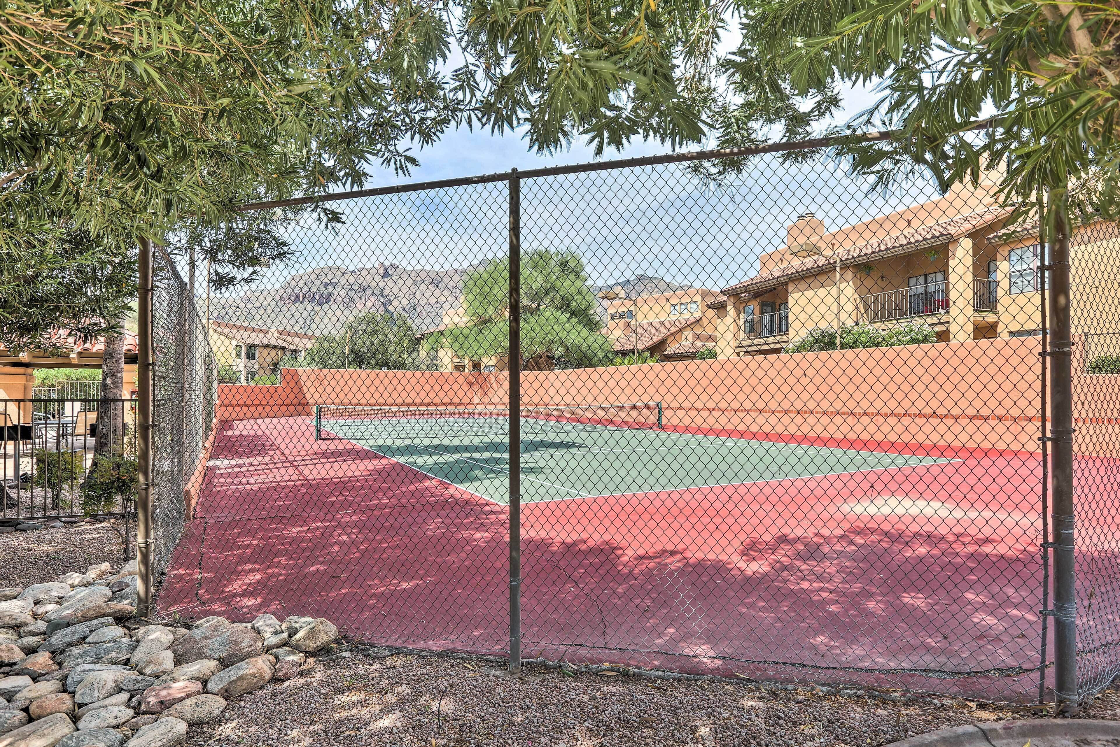 A fenced tennis court with a red surface, surrounded by a chain-link fence and trees.