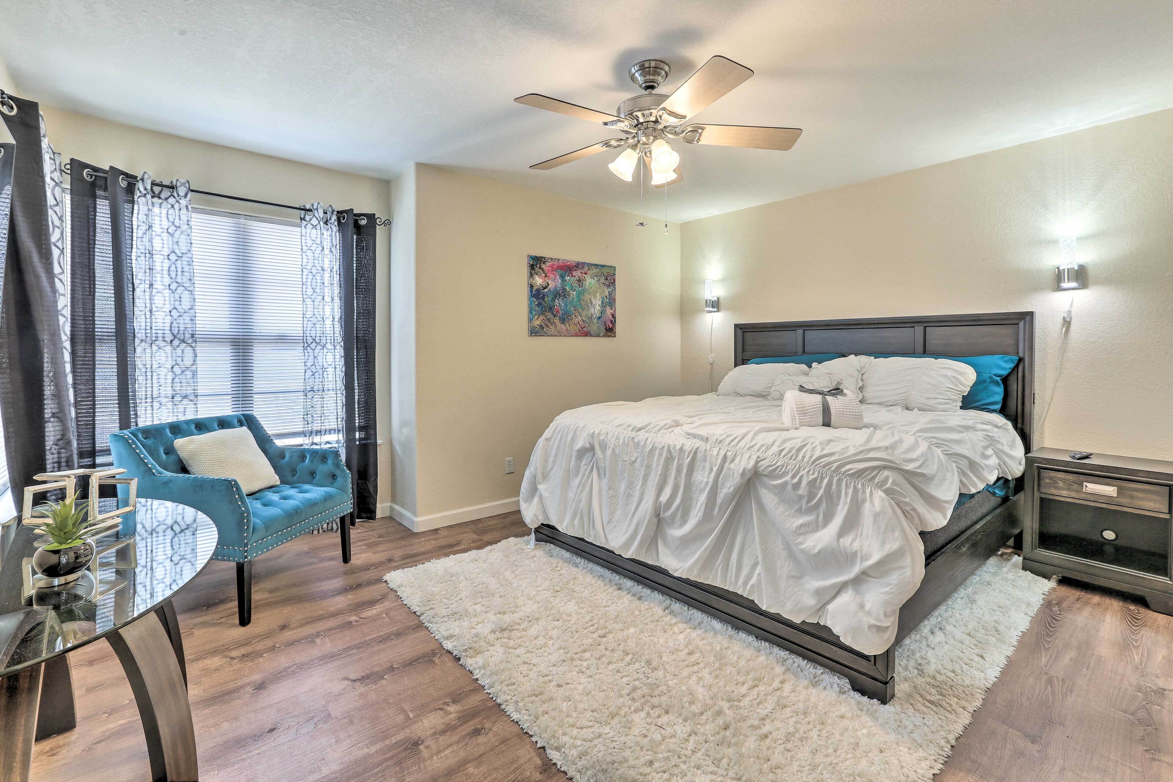 A bedroom with a bed, a blue armchair, a glass table, a ceiling fan, and a painting on the wall.