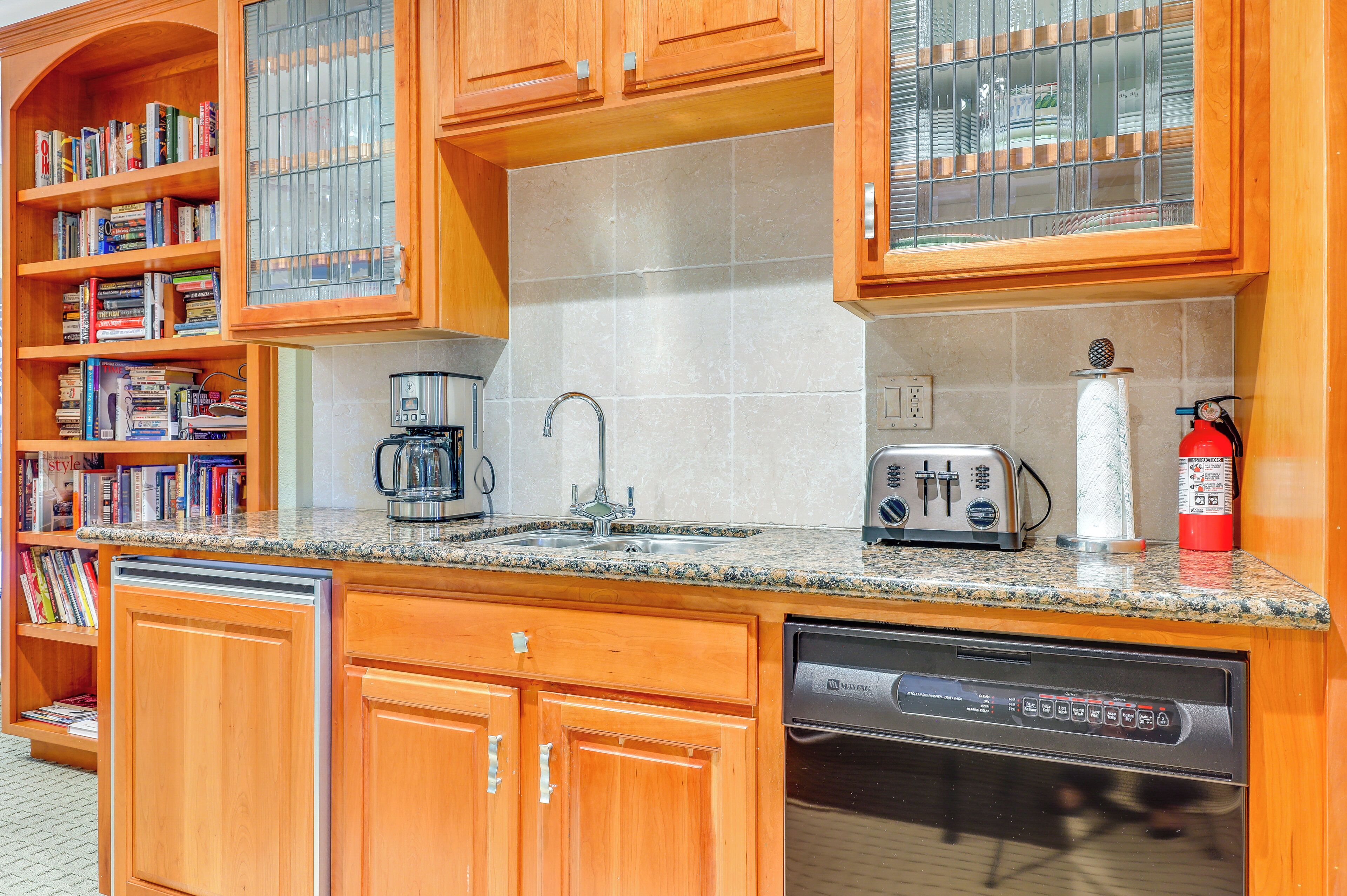 A kitchen with wooden cabinets, a granite countertop, a dishwasher, a coffee maker, a toaster, and a fire extinguisher.
