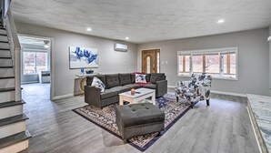 A spacious living room with a gray sectional sofa, a white coffee table, and a patterned rug. There is a staircase on the left and a door leading to another room. - Whitesburg Vacation Home w/ Glass-enclosed Porch (Whitesburg)